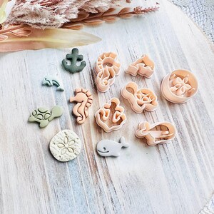 May include: A selection of sea-themed cookie cutters, featuring an anchor, seahorse, turtle, and whale designs. The cutters are in shades of green, beige, and light pink, displayed on a light wooden surface.