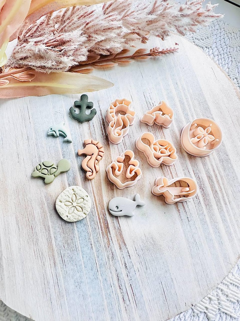 May include: A selection of sea-themed cookie cutters, featuring an anchor, seahorse, turtle, and whale designs. The cutters are in shades of green, beige, and light pink, displayed on a light wooden surface.