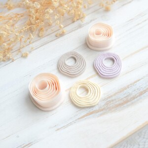 May include: A collection of handmade clay donut-shaped ornaments in various pastel colors, including pink, beige, gray, yellow, and lavender. The ornaments are arranged on a white wooden surface, with dried floral accents in the background.