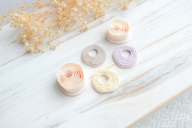 May include: A collection of handmade clay donut-shaped ornaments in various pastel colors, including pink, beige, gray, yellow, and lavender. The ornaments are arranged on a white wooden surface, with dried floral accents in the background.