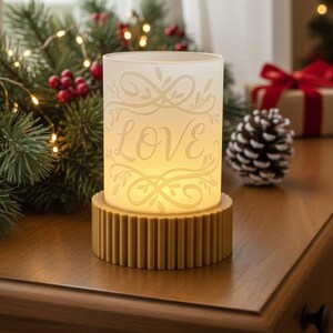 May include: A lit candle holder with the word "LOVE" etched into the frosted glass. The candle holder sits on a gold-coloured, ridged base. The background includes Christmas decorations.
