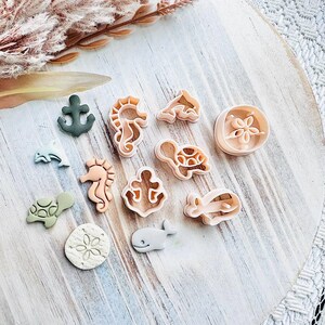 May include: A selection of sea-themed biscuit cutters, featuring a seahorse, turtle, whale, and anchor. The cutters are in beige, green, and grey, arranged on a light wooden surface. A sprig of dried flowers is in the background.