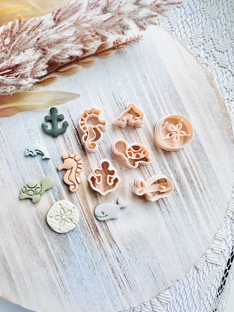 May include: A selection of sea-themed biscuit cutters, featuring a seahorse, turtle, whale, and anchor. The cutters are in beige, green, and grey, arranged on a light wooden surface. A sprig of dried flowers is in the background.