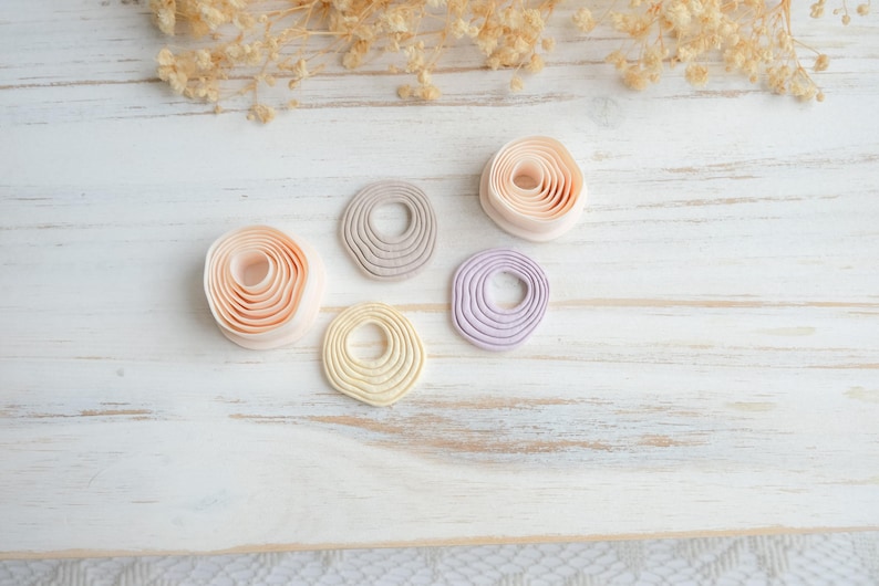 May include: Five handmade clay rings in soft pastel colors: peach, gray, yellow, and lavender. The rings have a layered, textured design and are arranged on a white wooden surface with dried flowers.
