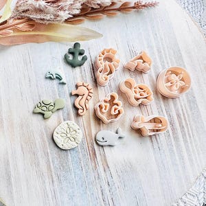 May include: A selection of sea-themed biscuit cutters, featuring a seahorse, whale, and anchor. The cutters are in shades of green, beige, and peach, arranged on a white wooden surface. The overall theme is nautical.
