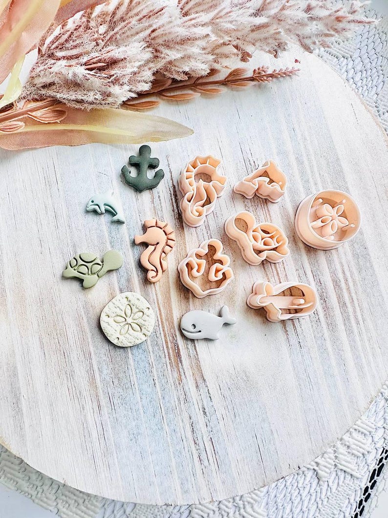 May include: A selection of sea-themed biscuit cutters, featuring a seahorse, whale, and anchor. The cutters are in shades of green, beige, and peach, arranged on a white wooden surface. The overall theme is nautical.