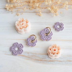May include: An assortment of flower-shaped cookie cutters and phone grips in shades of pink and purple, arranged on a white wooden surface. The cutters have detailed petal designs, and the phone grips feature gold rings.