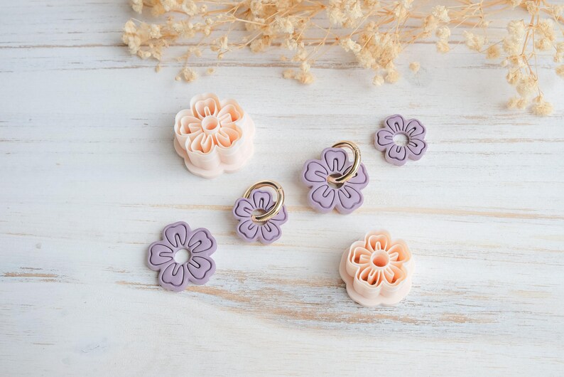 May include: An assortment of flower-shaped cookie cutters and phone grips in shades of pink and purple, arranged on a white wooden surface. The cutters have detailed petal designs, and the phone grips feature gold rings.