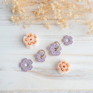 May include: An assortment of flower-shaped accessories in shades of peach and lavender, some with gold-colored rings. The items are arranged on a white, weathered wooden surface, with dried floral accents in the background.