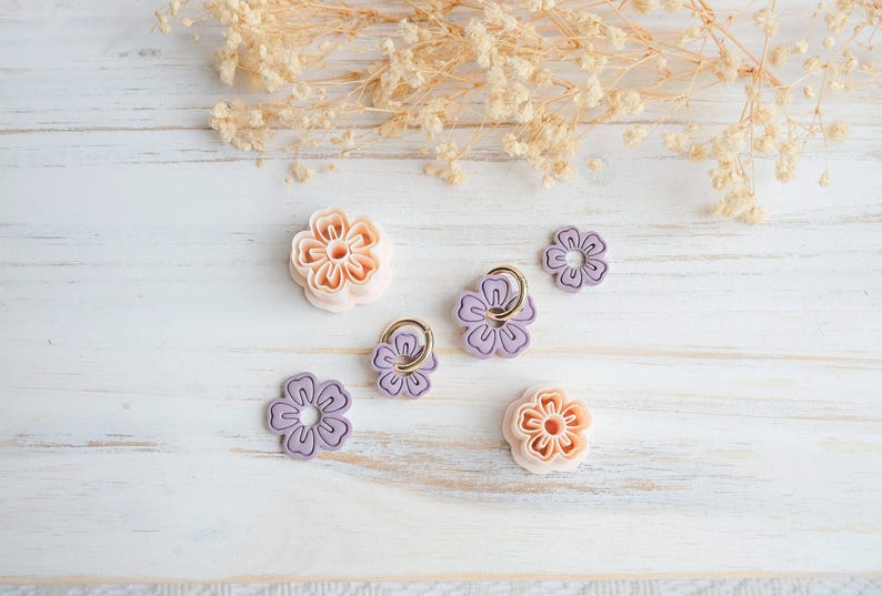 May include: An assortment of flower-shaped accessories in shades of peach and lavender, some with gold-colored rings. The items are arranged on a white, weathered wooden surface, with dried floral accents in the background.