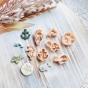 May include: A selection of sea-themed biscuit cutters, featuring an anchor, seahorse, turtle, and whale. The cutters are in beige, green, and grey, arranged on a light wooden board. A sprig of dried flowers is in the background.