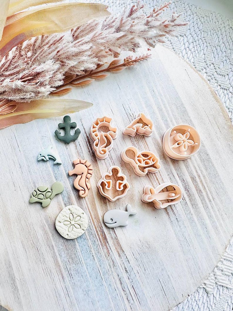 May include: A selection of sea-themed biscuit cutters, featuring an anchor, seahorse, turtle, and whale. The cutters are in beige, green, and grey, arranged on a light wooden board. A sprig of dried flowers is in the background.