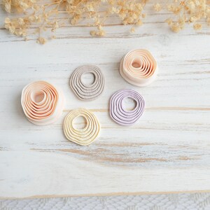 May include: Five handmade clay pieces in shades of peach, gray, yellow, and lavender, arranged on a white wooden surface. Each piece has a circular shape with concentric ridges, resembling a coiled design. Dried floral accents are in the background.