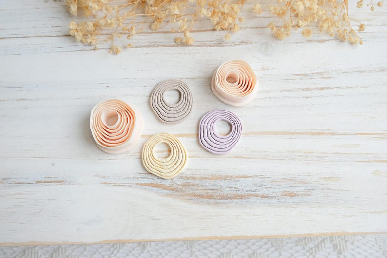 May include: Five handmade clay pieces in shades of peach, gray, yellow, and lavender, arranged on a white wooden surface. Each piece has a circular shape with concentric ridges, resembling a coiled design. Dried floral accents are in the background.