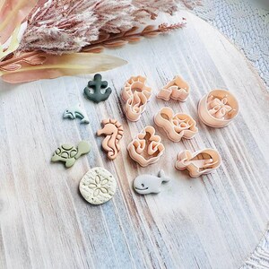 May include: A selection of sea-themed cookie cutters in shapes like a seahorse, whale, and anchor. The cutters are in pink, green, and grey, arranged on a light wooden surface.