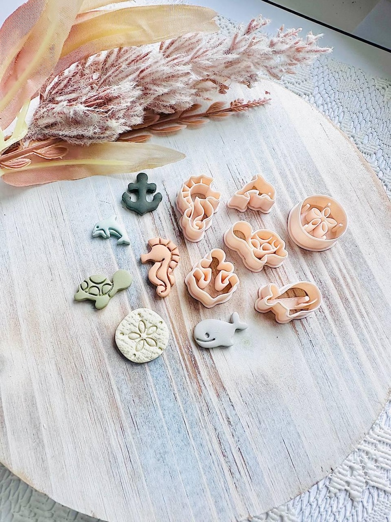May include: A selection of sea-themed cookie cutters in shapes like a seahorse, whale, and anchor. The cutters are in pink, green, and grey, arranged on a light wooden surface.