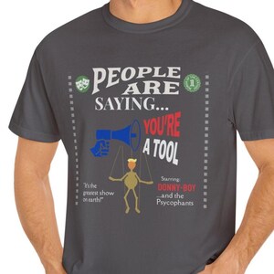 Puede incluir: Una camiseta gris oscuro con un estampado gráfico de un títere con un megáfono que dice "You're a Tool". El gráfico también incluye el texto "People are saying..." y "It's the greatest show on earth!" y "Starring: Donny-Boy...and the Psycophants".