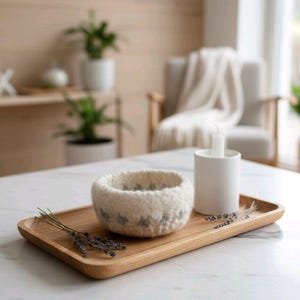 Könnte beinhalten: Ein Holztablett mit einer weißen Filzschale mit grauen Sternmustern, einem weißen Kerzenhalter mit einer brennenden Kerze und Lavendelzweigen. Das Tablett steht auf einer weißen Marmoroberfläche, mit einem neutralen Hintergrund.