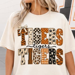 May include: A cream-colored short-sleeve t-shirt featuring a graphic design with the word TIGERS repeated in two rows, separated by the word tigers in script. The letters feature various patterns including tiger stripes, stars, and polka dots.