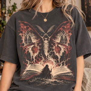 May include: A charcoal gray short-sleeve t-shirt featuring a large graphic print of a moth with gothic arched windows on its wings, positioned above a figure kneeling before an open, glowing book with visible text on the pages.