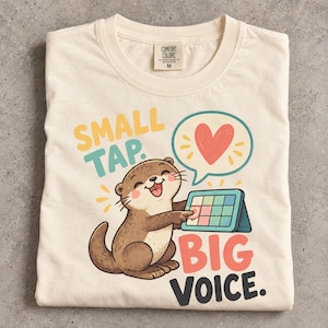 May include: A cream-colored t-shirt featuring a graphic of a brown otter tapping a tablet screen. The text reads SMALL TAP. BIG VOICE. A speech bubble above the otter contains a red heart. The shirt has a Comfort Colors brand label.