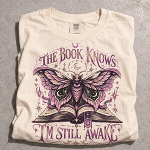 May include: A cream-colored t-shirt featuring a graphic of a moth with purple and black wings resting on an open book. The text reads THE BOOK KNOWS and I'M STILL AWAKE. The design includes celestial elements like stars and crescent moons.