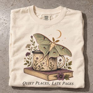 May include: A cream-colored t-shirt featuring a graphic of a large green moth perched on a speckled mug, which rests on a closed book. The design includes small purple and white flowers, a crescent moon, and the text "QUIET PLACES, LATE PAGES."
