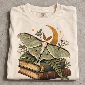 May include: A cream-colored t-shirt featuring a graphic print of a large pale green moth resting on a stack of two vintage-style books. The design includes a golden crescent moon, small stars, fern leaves, and white flowers. The shirt has a Comfort Colors label.