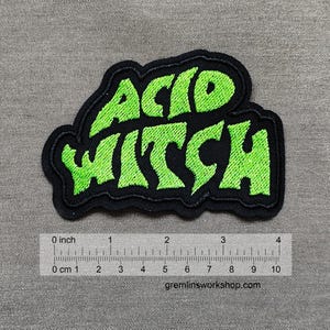 Op de afbeelding: Een zwarte en groene geborduurde patch met de tekst "ACID WITCH" in een gestileerd lettertype.