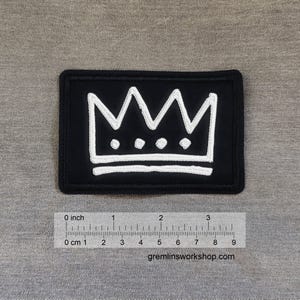 May include: Black rectangular patch featuring a white crown design. The crown has a jagged top with three circular dots below. The patch is approximately 3 inches long. The website gremlinsworkshop.com is printed at the bottom.