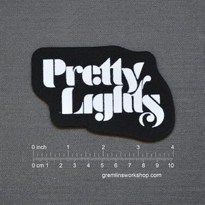 May include: Black patch with the words "Pretty Lights" in white, stylized lettering. The patch is approximately 10 cm wide and has a unique, irregular shape. The website gremlinsworkshop.com is printed at the bottom.