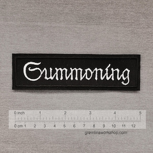 Puede incluir: Un parche rectangular negro bordado con el texto blanco "Summoning" en una fuente estilizada. Una regla de plástico transparente colocada debajo del parche muestra que mide aproximadamente 11,5 cm de largo y 2,5 cm de alto.