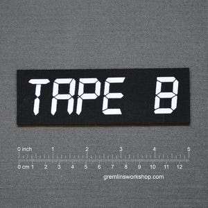 Op de afbeelding: Zwart rechthoekig embleem met de tekst "TAPE B" in witte digitale lettertype. Een liniaal met inches en centimeters staat onder het embleem. De website gremlinsworkshop.com staat onderaan.