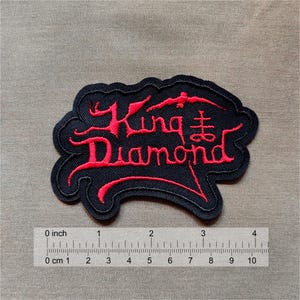 May include: A black embroidered patch featuring the red text King Diamond and a sulfur cross symbol. The patch is positioned above a ruler showing a width of approximately 3.5 inches and a height of approximately 2.25 inches for the design.