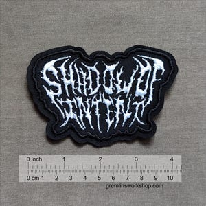 Pode incluir: Etiqueta bordada preta com letras brancas que dizem "SHADOW OF INTENSITY".