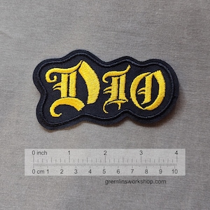 May include: Embroidered patch featuring the word "DIO" in yellow gothic lettering on a navy blue background. The patch is approximately 10 cm long and has a wavy border. The website gremlinsworkshop.com is visible at the bottom.