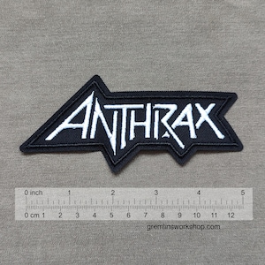May include: Black and white embroidered patch featuring the band name "ANTHRAX" in a stylized font. The patch is approximately 4 inches wide and has a unique arrow-like shape. The background is a neutral gray fabric.