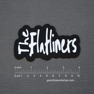 Puede incluir: Un parche negro con el texto blanco "The Flatliners" en una fuente estilizada. El parche mide aproximadamente 7,6 cm de largo y 3,8 cm de alto. El sitio web gremlinsworkshop.com está impreso en la parte inferior.