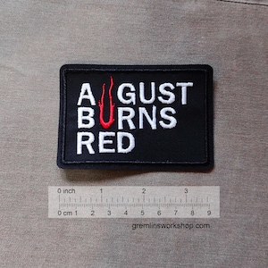 以下が含まれることがあります： 黒色の刺繍パッチ。白い文字で「AUGUST BURNS RED」と書かれ、赤い炎のデザインが施されています。パッチは長方形で、幅は約7.6cmです。タン色の生地の背景に配置されています。
