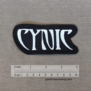Puede incluir: Parche bordado negro con la palabra "CYNIC" en letras blancas. El parche mide aproximadamente 5 cm de ancho y 2,5 cm de alto.