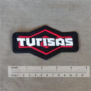 May include: A black and red TUTISAS patch with white lettering. The patch is rectangular with a diamond-shaped design in the center. The patch is approximately 4 inches long.