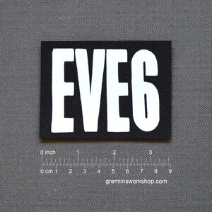 May include: A black rectangular patch featuring the word "EVE6" in bold white letters. The patch is on a grey surface with a ruler showing centimetres and inches. The website gremlinsworkshop.com is visible.