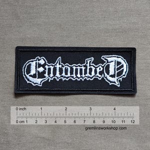 May include: Black embroidered patch with the white text "Entombe D" in a gothic font style. The patch is approximately 4 inches wide.