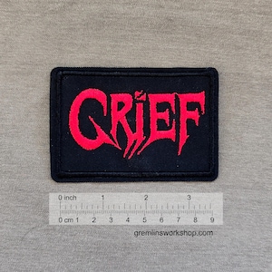 Pode incluir: Patch retangular preto com a palavra "GRIEF" bordada em vermelho, com letras estilizadas. O patch tem aproximadamente 7.6 cm de largura. O site gremlinsworkshop.com está impresso na parte inferior.