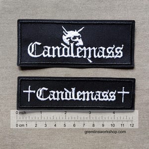 May include: Two black rectangular patches with white embroidered text that reads "Candlemass". One patch features a skull design above the text. The other patch has a cross on each end. A ruler shows the patches are approximately 4 inches long.
