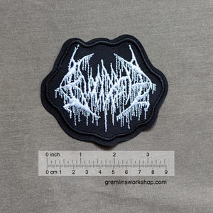 Op de afbeelding: Zwarte geborduurde patch met een wit bandlogo met de tekst "SUFFOCATE". De patch is ongeveer 5 cm in diameter.