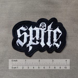 May include: Black and white embroidered patch with the word "spite" in a gothic font. A small skull with a pentagram is above the word.