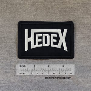Könnte beinhalten: Schwarzer rechteckiger Aufnäher mit dem Wort "HEDEX" in Weiß gestickt. Der Aufnäher ist etwa 7,6 cm lang. Die Website gremlinsworkshop.com ist unten aufgedruckt.