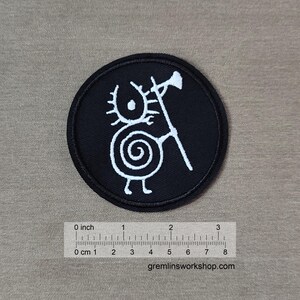 Puede incluir: Un parche redondo negro con un diseño de criatura blanca estilizada. La criatura tiene un cuerpo en espiral, dos piernas y una cabeza con un diseño solar. Sostiene un bastón con una bandera. El parche mide aproximadamente 7.6 cm de diámetro. El sitio web gremlinsworkshop.com es visible.
