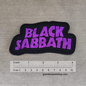 May include: A rectangular, embroidered patch with the words "BLACK SABBATH" in purple, stylized lettering. The patch has a black border and is approximately 4 inches long. The background is a neutral gray fabric.